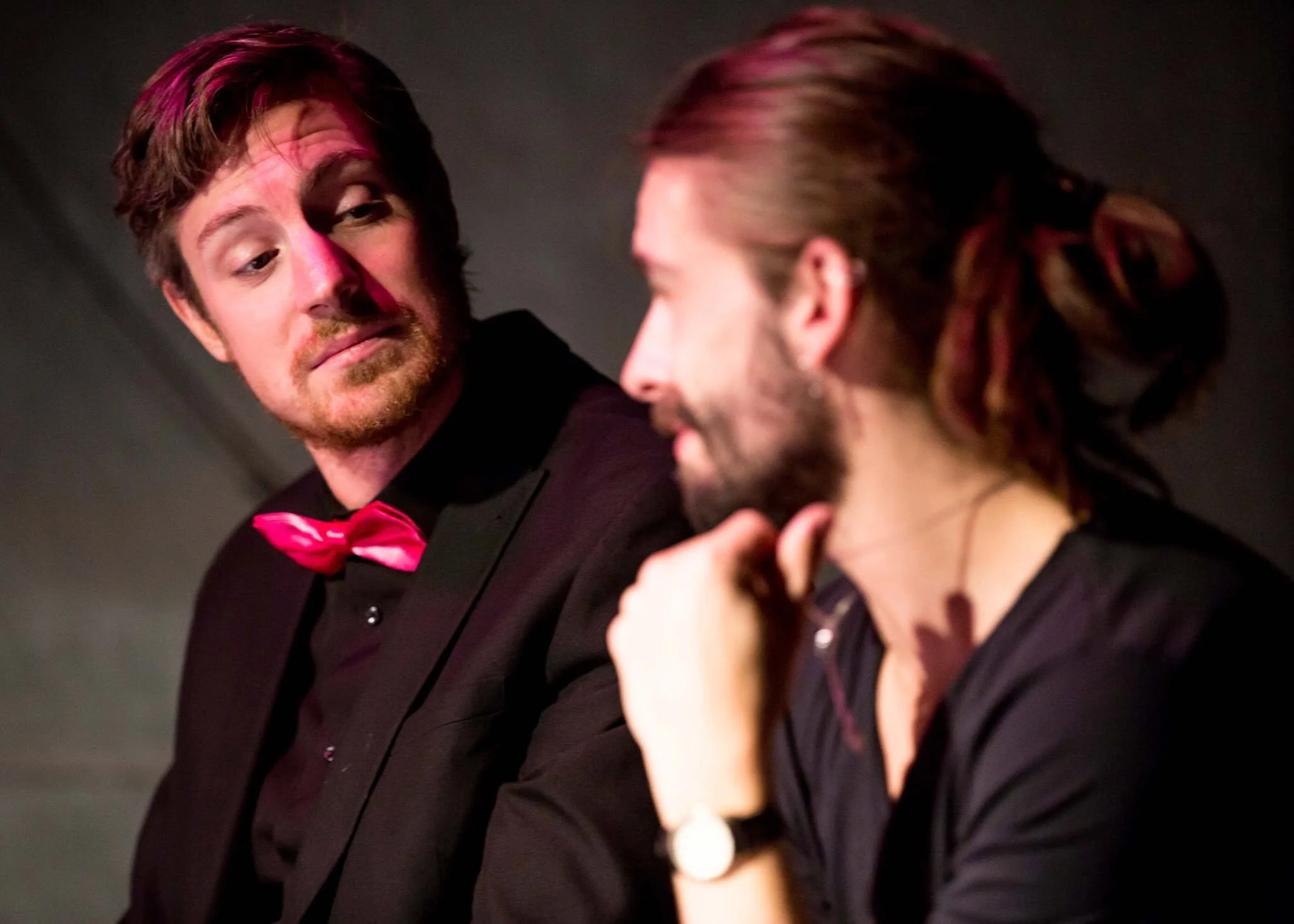 Two men dressed in formal black attire, with one wearing a pink bow tie, engage in conversation against a dark background. One man has light brown hair with a slight wave, the other has long, dark hair tied back.