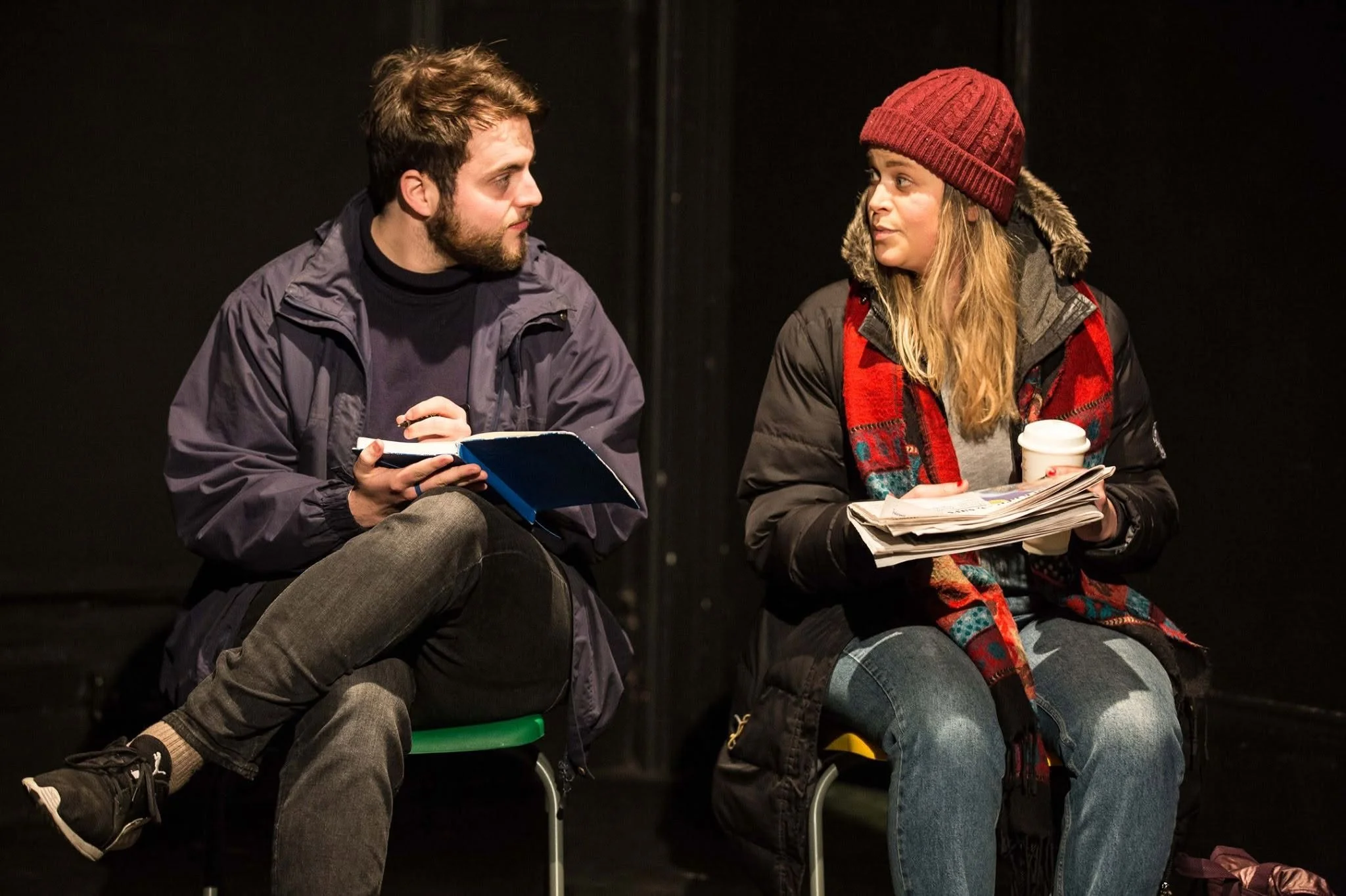 Two people sitting on chairs and engaged in conversation, with theatre or stage setting background. The man on the left has a beard, dark hair, and is wearing a jacket, and is holding a blue notebook. The woman on the right has long blonde hair, is w