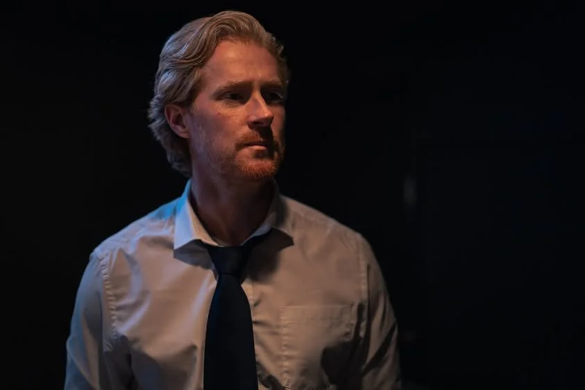 A man with blond hair and a beard wearing a white shirt and dark tie, looking serious in a dimly lit room.