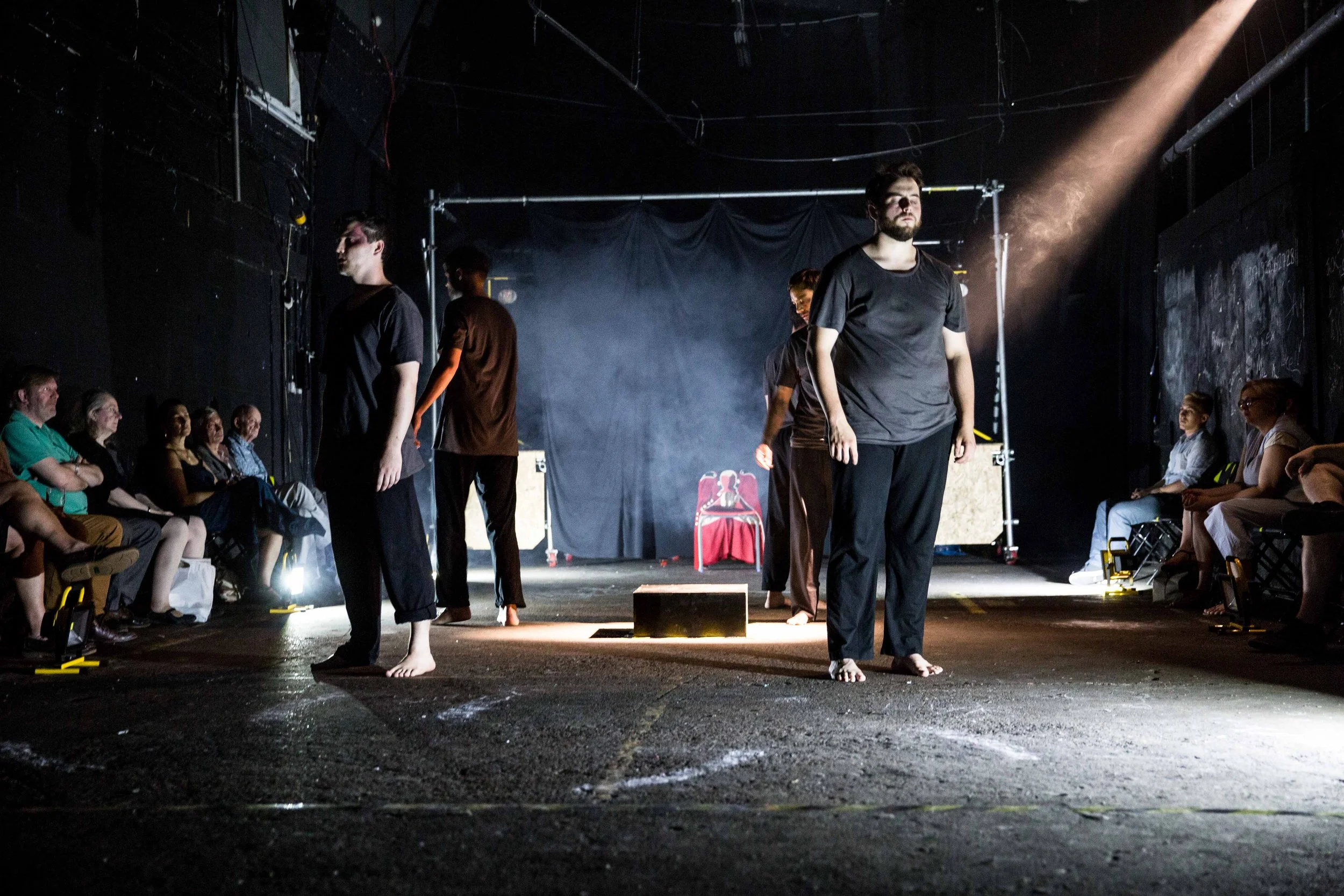 A group of people performing a contemporary theatre piece on a dark stage with dramatic lighting, surrounded by an audience seated on each side.