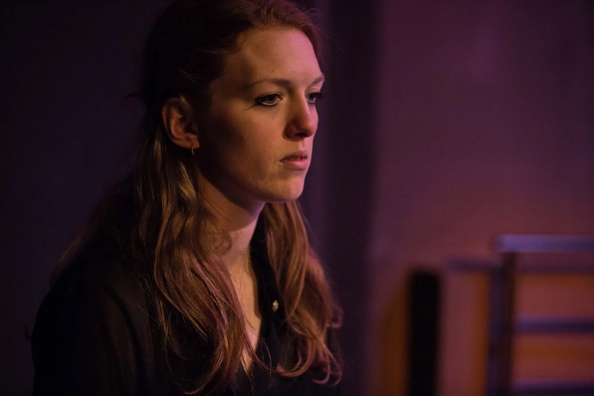 A young woman with long wavy hair looking down thoughtfully in a dimly lit room with purple and pink lighting.