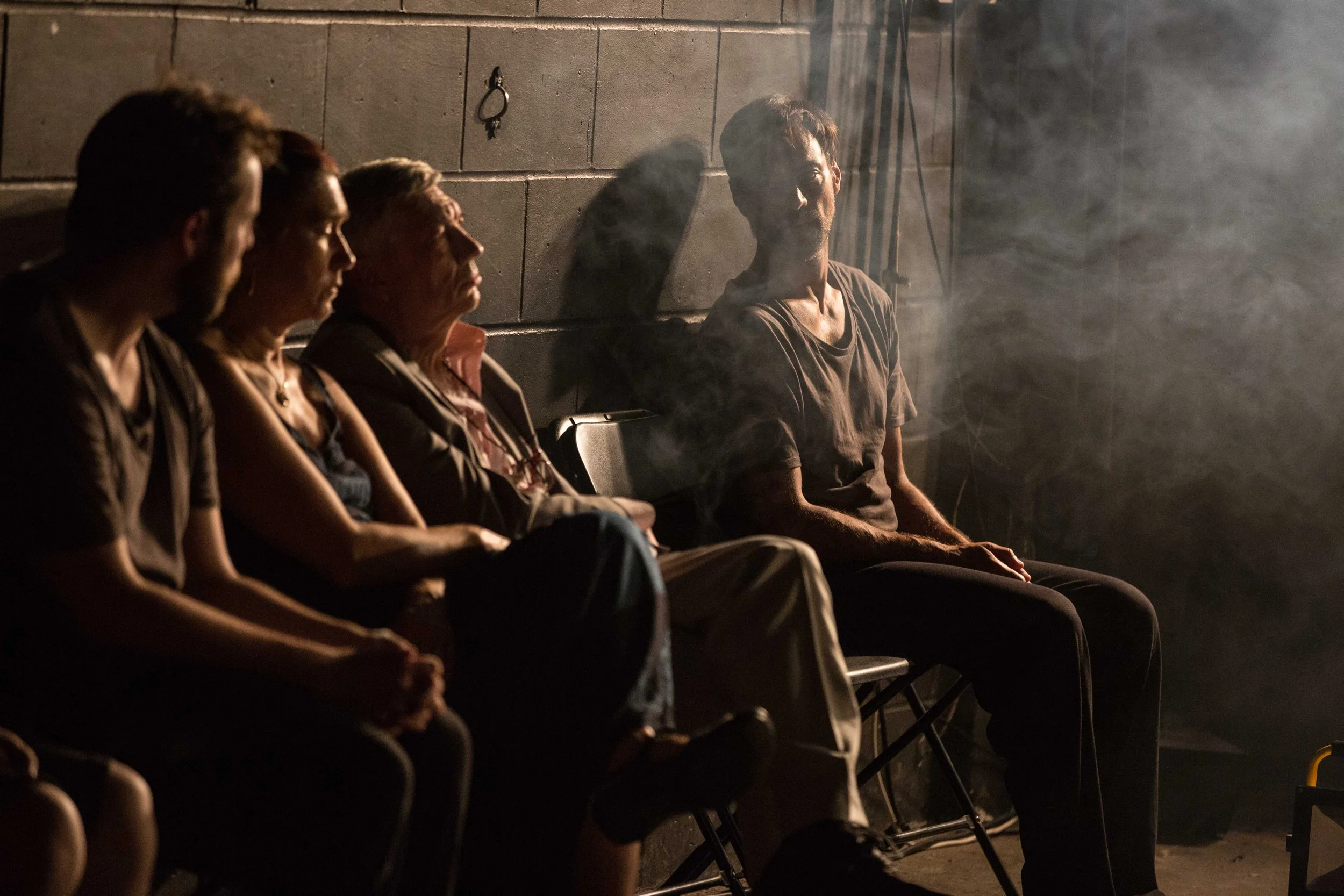 Four people are sitting on chairs against a concrete wall, with one person on the right shaded and looking directly at the camera, while the others are in profile or facing away, in a dim, smoky environment.