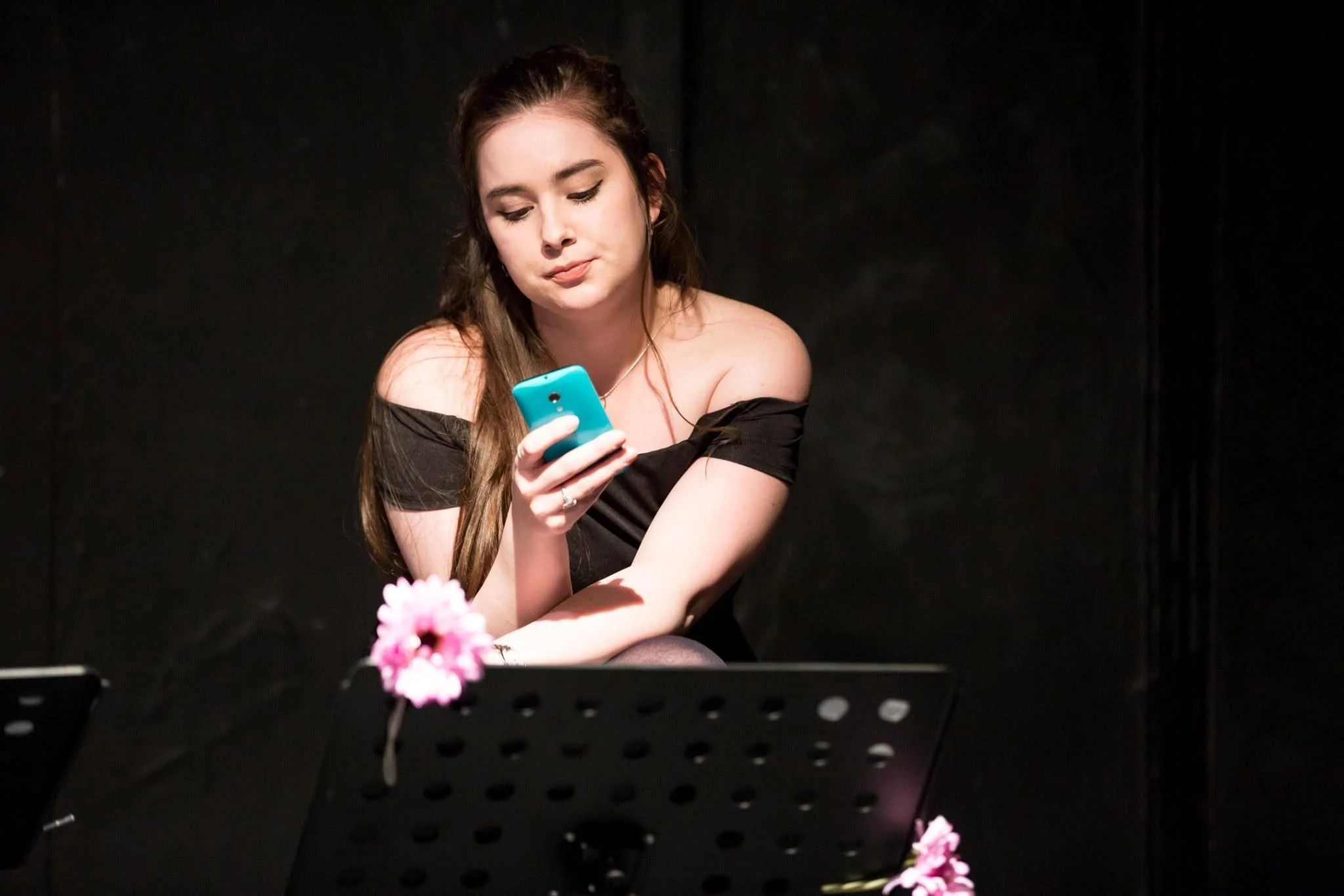 Young woman with long hair, wearing a black off-shoulder top, looking at her phone, sitting at a table with pink flowers, against a dark background.