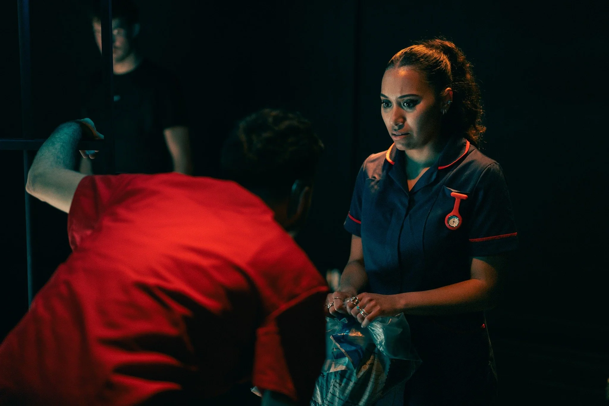 A woman in a dark medical uniform with red accents and a stethoscope appears concerned as she interacts with a person wearing a red shirt, whose back is to the camera, in a dimly lit room.