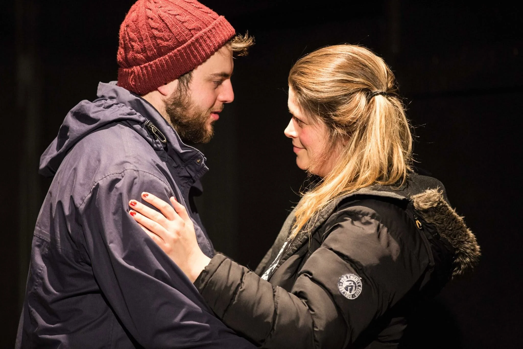 A man and woman face to face, touching each other's shoulders and smiling warmly at each other, set against a dark background.