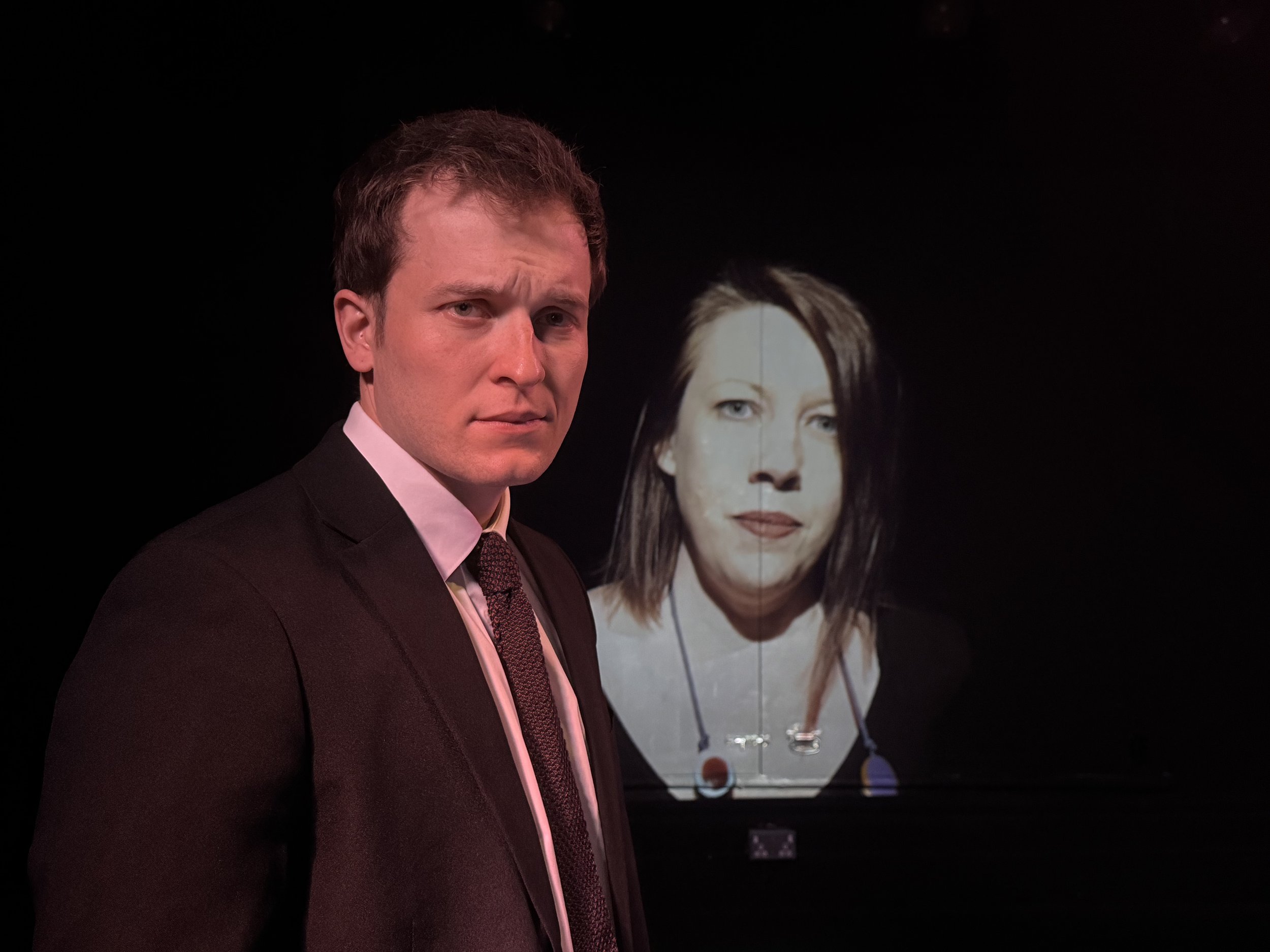 A man in formal attire standing in front of a display with a woman's portrait. The background is dark, highlighting the man and the portrait behind him.