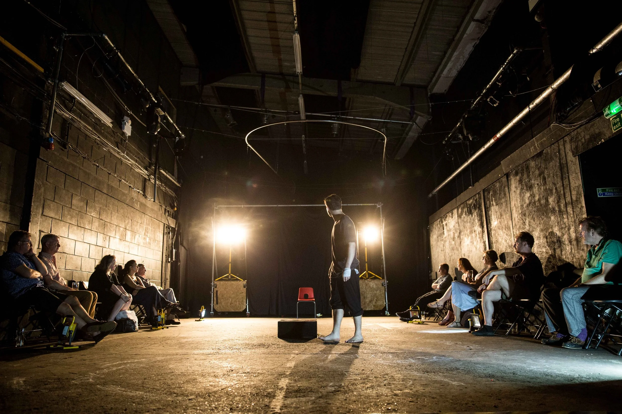 A person standing barefoot in the center of a dimly lit theater or performance space, facing away from the camera, while an audience sits on either side, watching. The room has exposed ceiling beams and industrial pipes, with bright lights illuminati