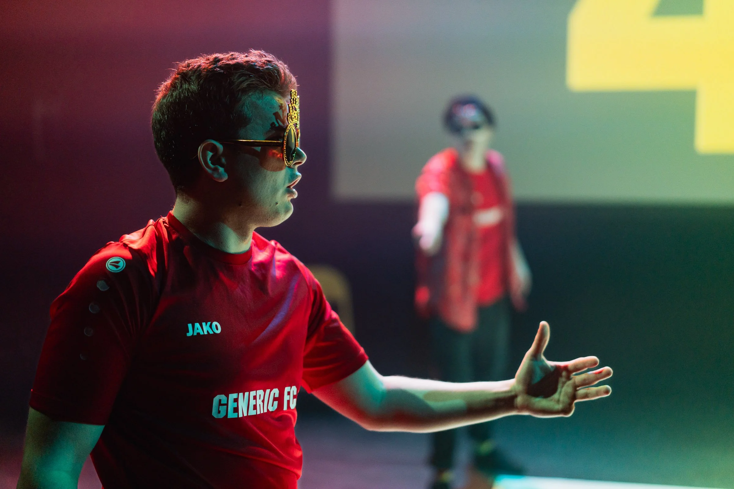 A young male athlete wearing sunglasses and a red sports shirt with 'JAKO' logo is in a sports hall, gesturing with his right hand while looking to the side. In the background, another person is visible, but blurred.