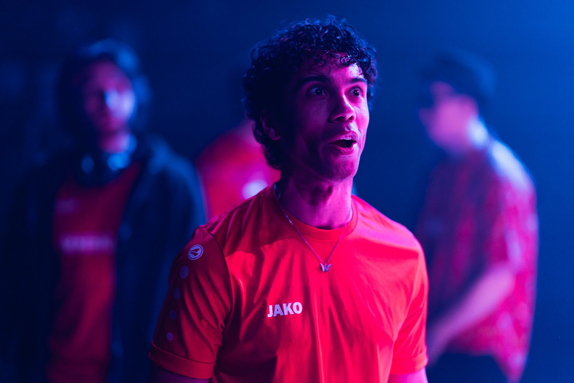 A man with curly hair wearing a red sports jersey with the brand name 'JAKO' on it, appearing surprised or excited in a neon-lit environment.