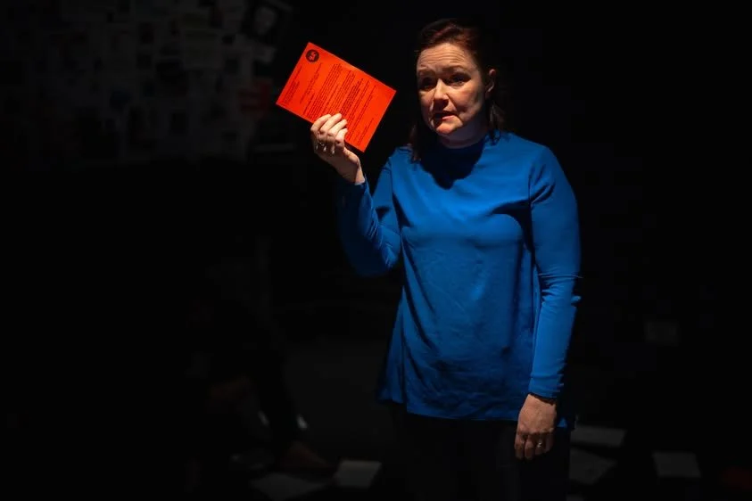 A woman in a blue top holding a bright red pamphlet in a dark room with a serious expression.
