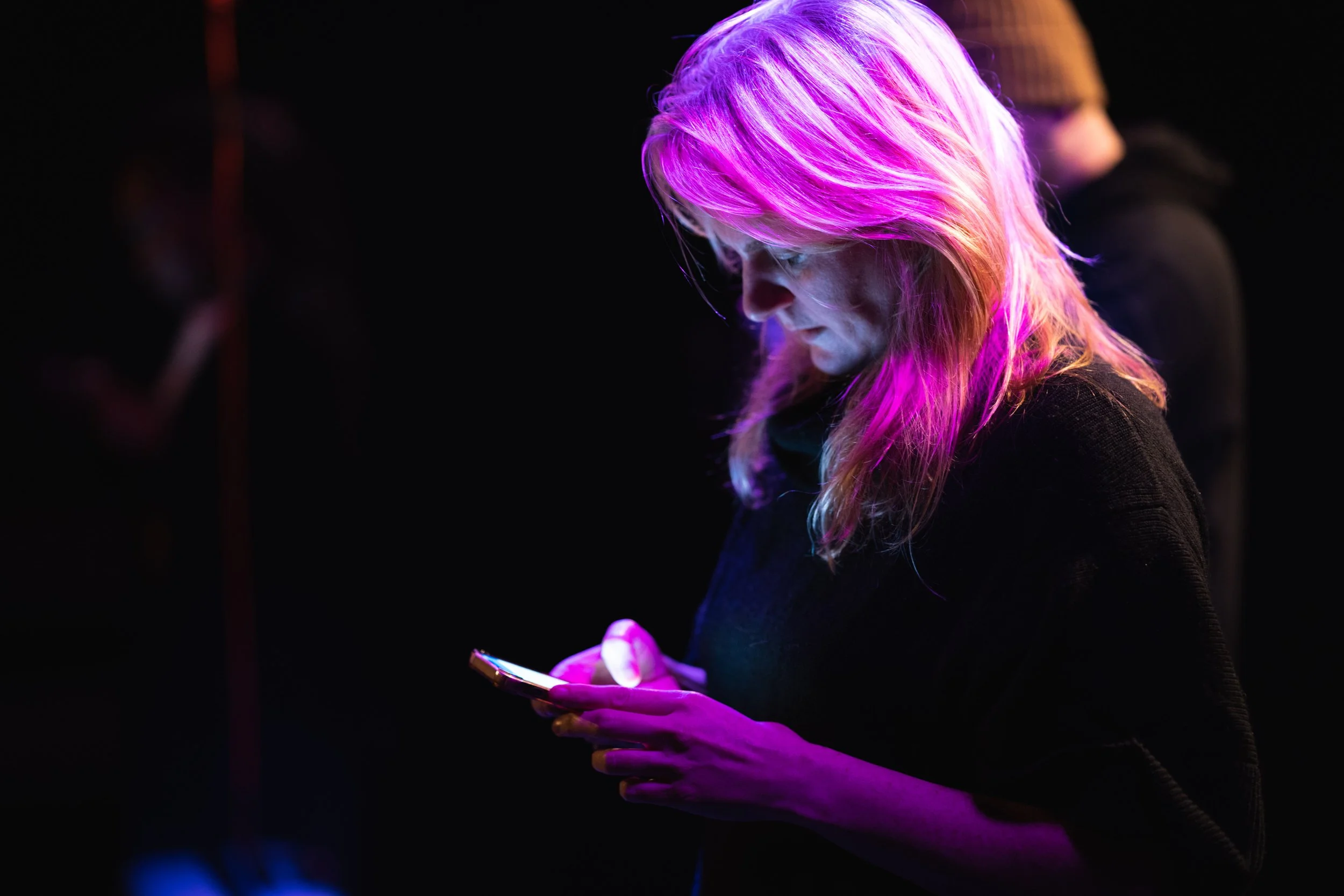A blonde woman lit in shades of pink and purple looking at her phone in a dark setting with neon lighting.