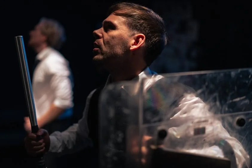 A man holding a glowing baton or light stick, with a woman blurred in the background, in a dark setting.