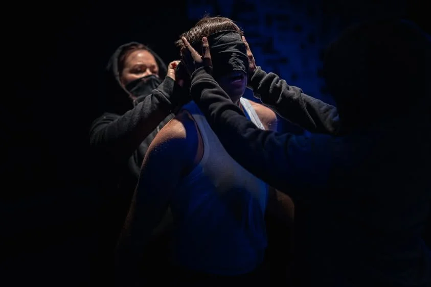A man wearing a white tank top is blindfolded as two people assist in adjusting the blindfold in a dark environment.
