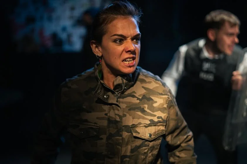 Woman with angry expression wearing camouflage jacket, in a heated scene with a man in background holding a riot shield.
