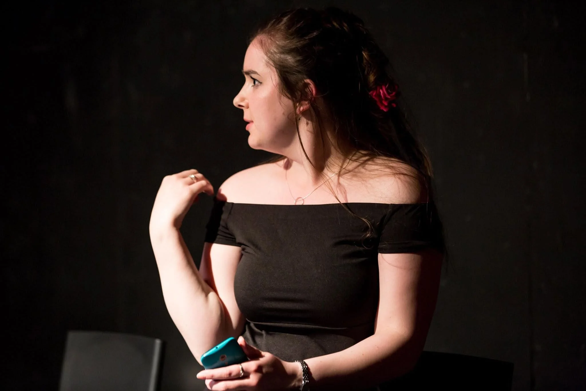 A woman with brown hair, wearing a black off-the-shoulder top, holds a phone in her left hand while looking to her right against a dark background.