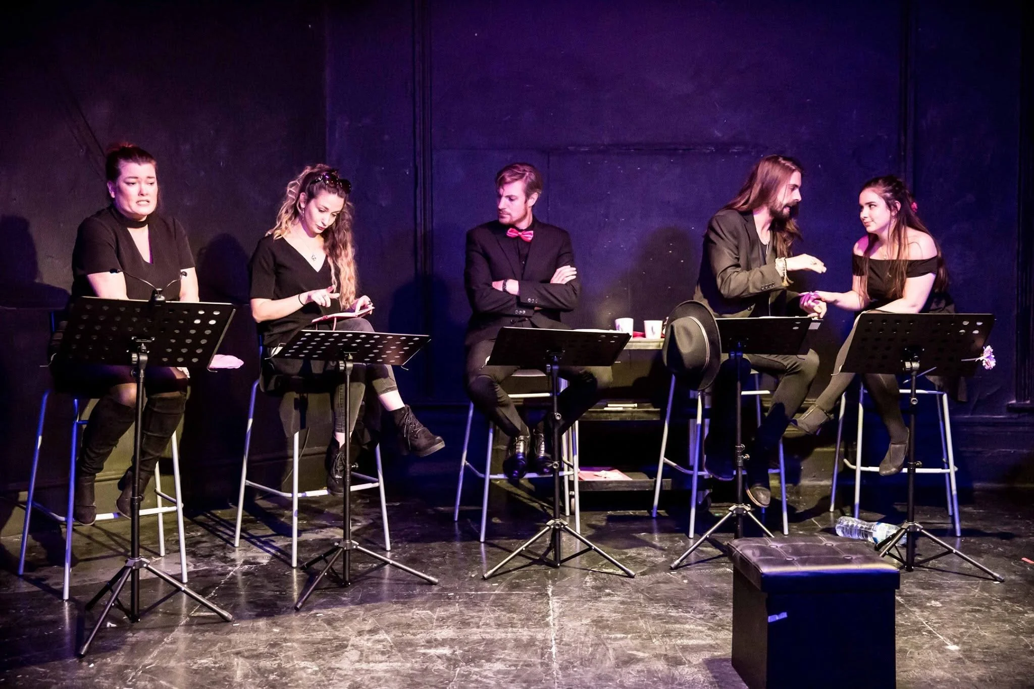 Six performers on a stage, some sitting, some standing, engaged in a theatrical performance, with music stands and a dark background surrounding them.