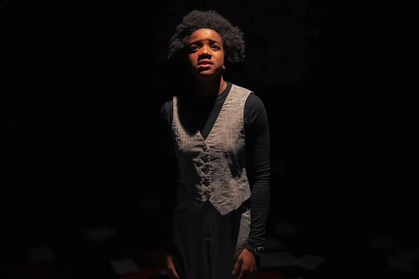 A young woman with natural afro hair stands in a dark room, illuminated by a single light source from above, casting shadows on her face and body.