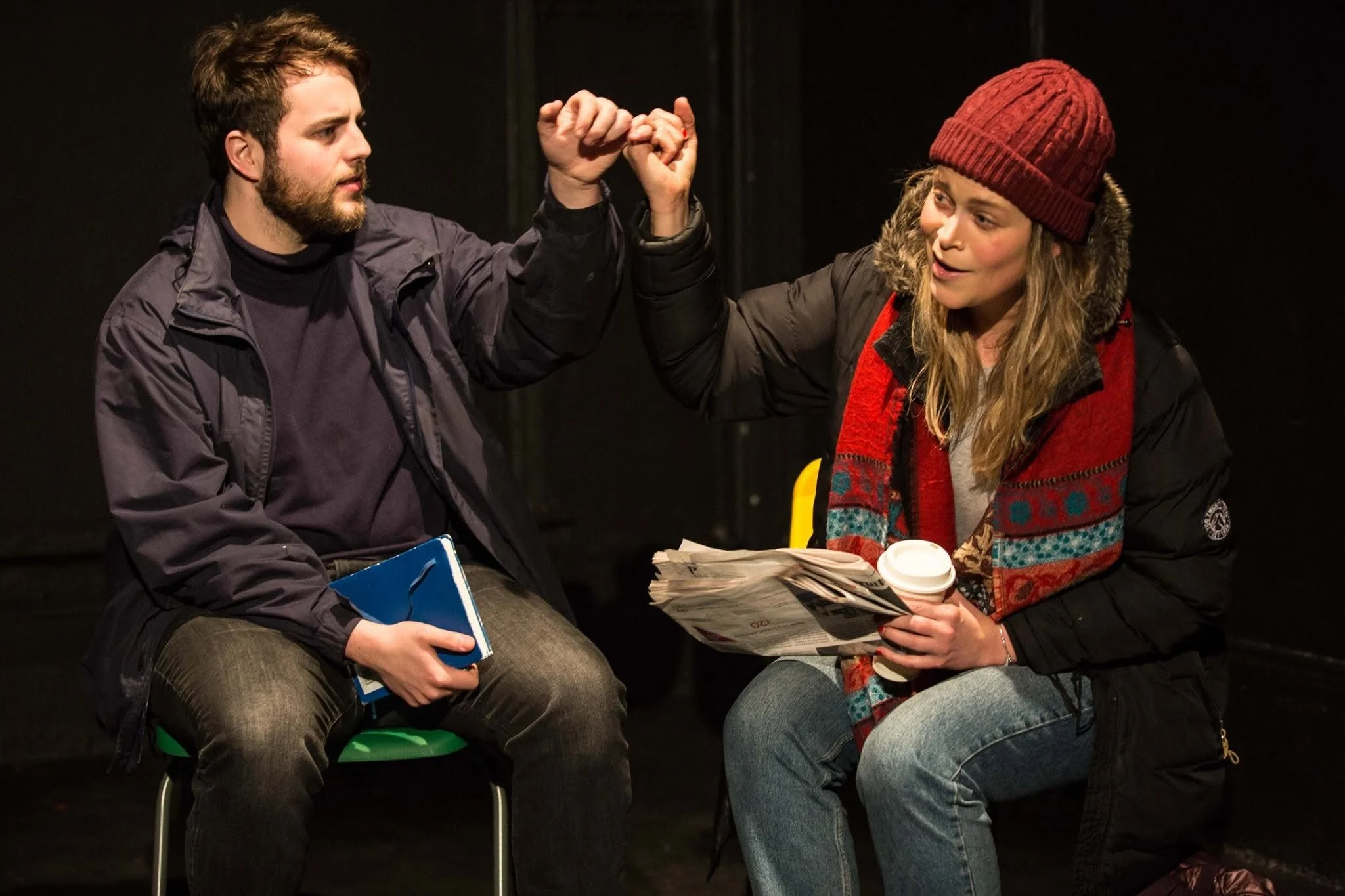 Two people sitting on chairs, engaging in friendly banter, with one person on the left holding a blue folder and the person on the right holding a cup of coffee, in a dark setting.