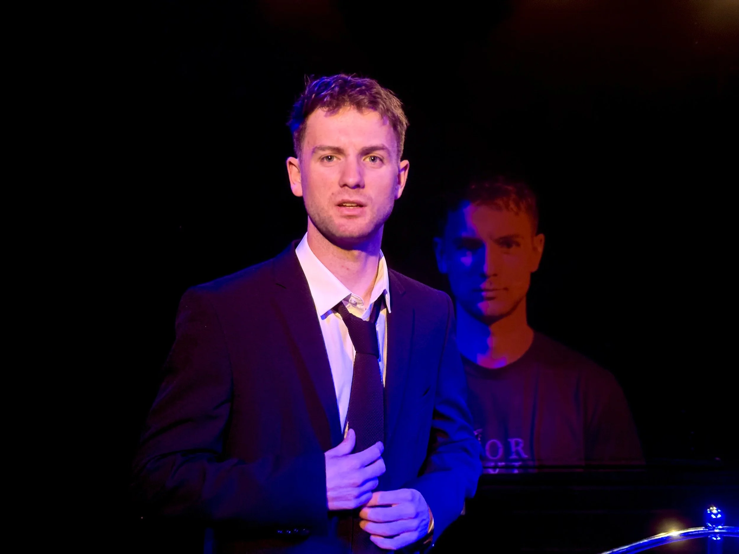 Young man in a suit speaking on stage, with another man partly visible in the background under stage lights.