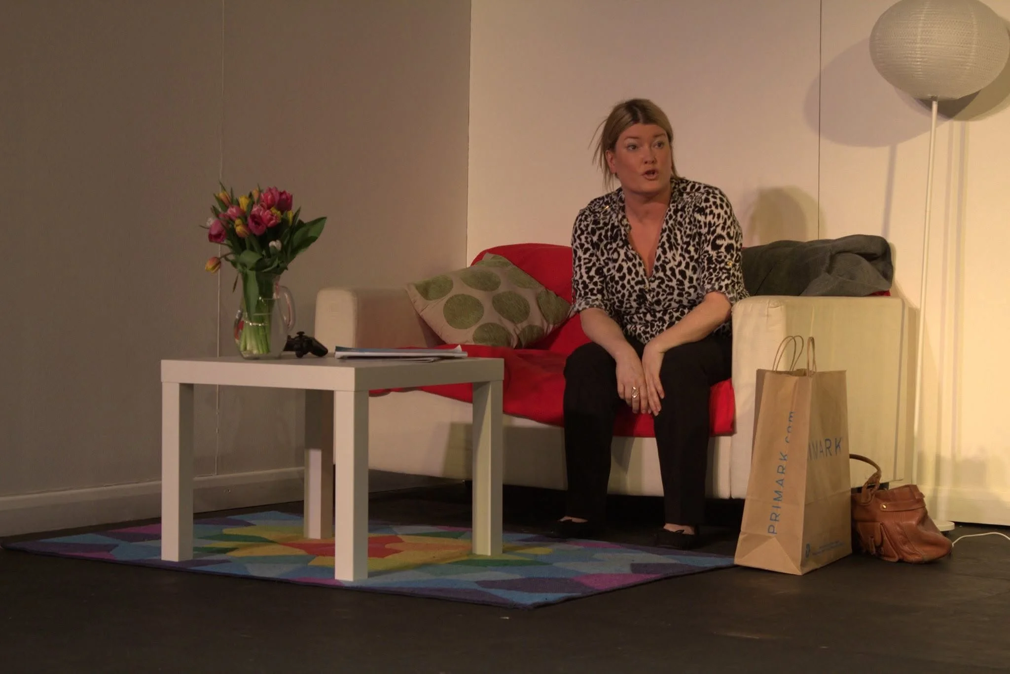 A woman sitting on a red couch with a leopard print top, talking. A white side table with a vase of pink and yellow flowers, some papers, and a game controller. Brown shopping bags and a brown handbag on the floor. A white lamp and gray wall in the b
