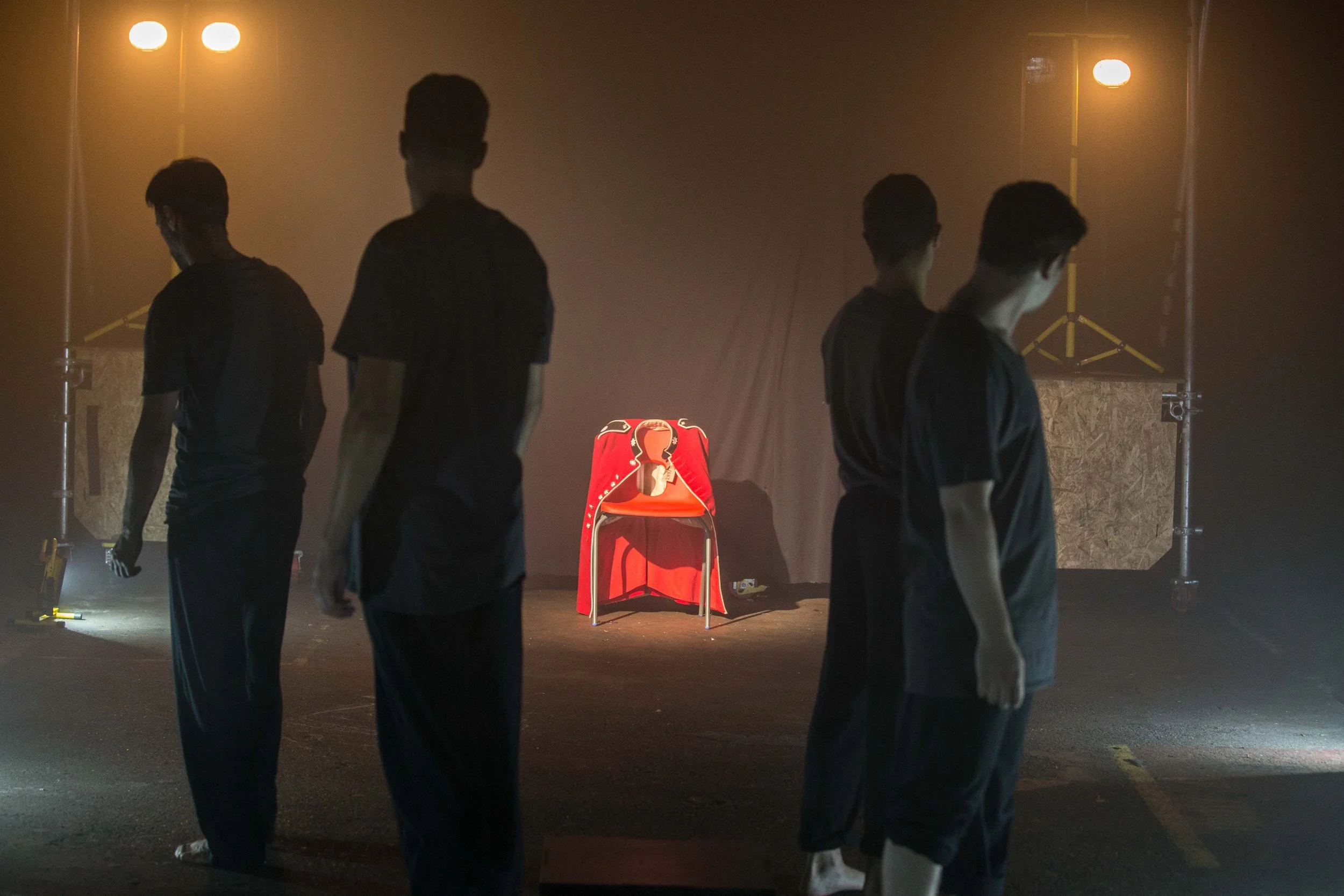 Group of four young men standing in a dimly lit room, facing away from the camera. In the background, there is a red chair with a red jacket and some stage lights.