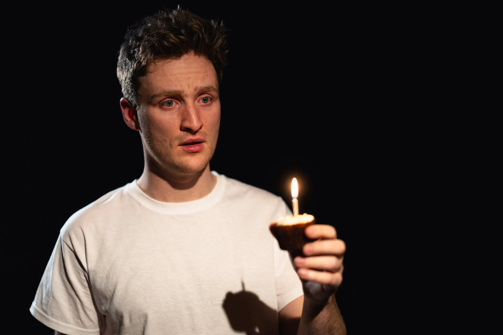 A young man with brown hair and wearing a white T-shirt is holding a cupcake with a lit birthday candle, looking at it with a surprised or thoughtful expression, against a black background.
