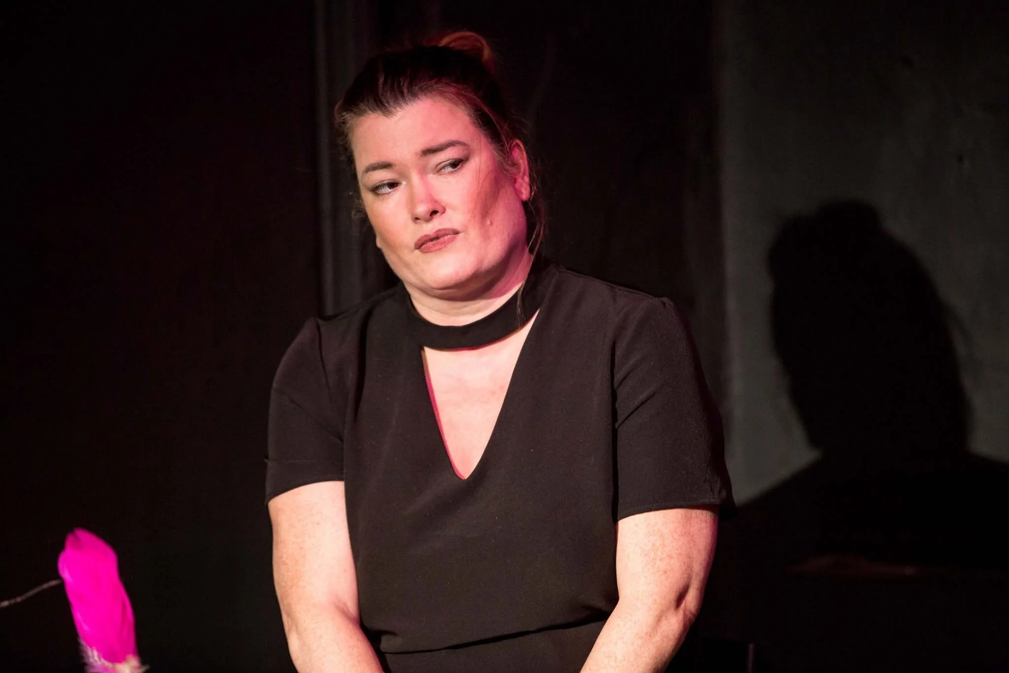 Woman with dark hair tied in a bun, wearing a black top with a scoop neck, looking to her right, sitting in a dark room.