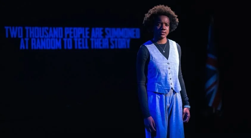 A woman on stage with a dark background, blue stage lighting, and text that reads 'Two thousand people are summoned at random to tell their story.' She has curly hair, wears a light-colored vest over a black shirt, with loose pants, and looks serious