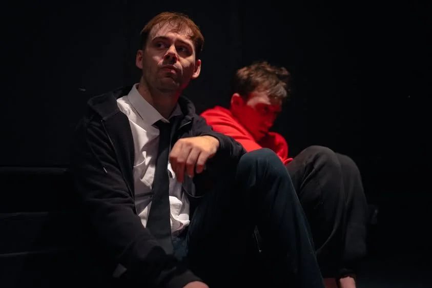 Two young men sitting in a dark room, one in a white shirt and black tie with a black jacket, looking thoughtfully into the distance, the other in a red hoodie looking down.