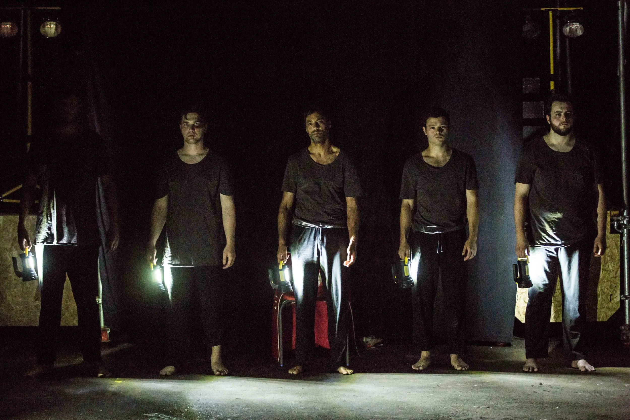 A group of six men dressed in black standing in a dark room with minimal lighting, some holding flashlights, facing forward.