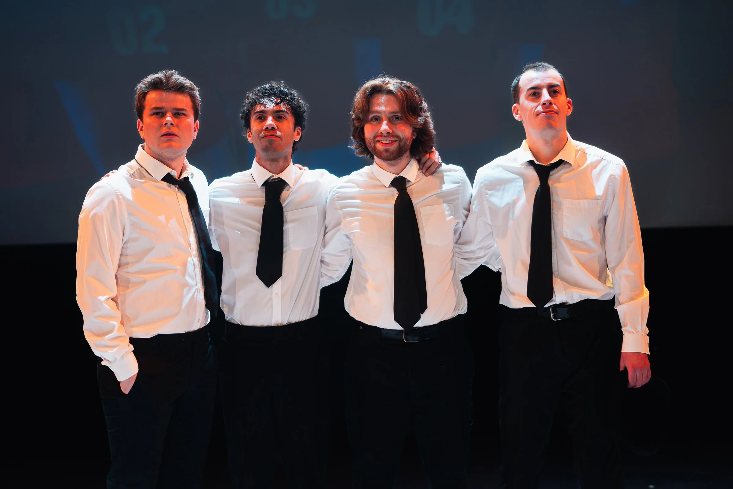 A group of four men dressed in white shirts and black ties standing closely together on a dark stage. They appear to be looking in different directions and have serious expressions.