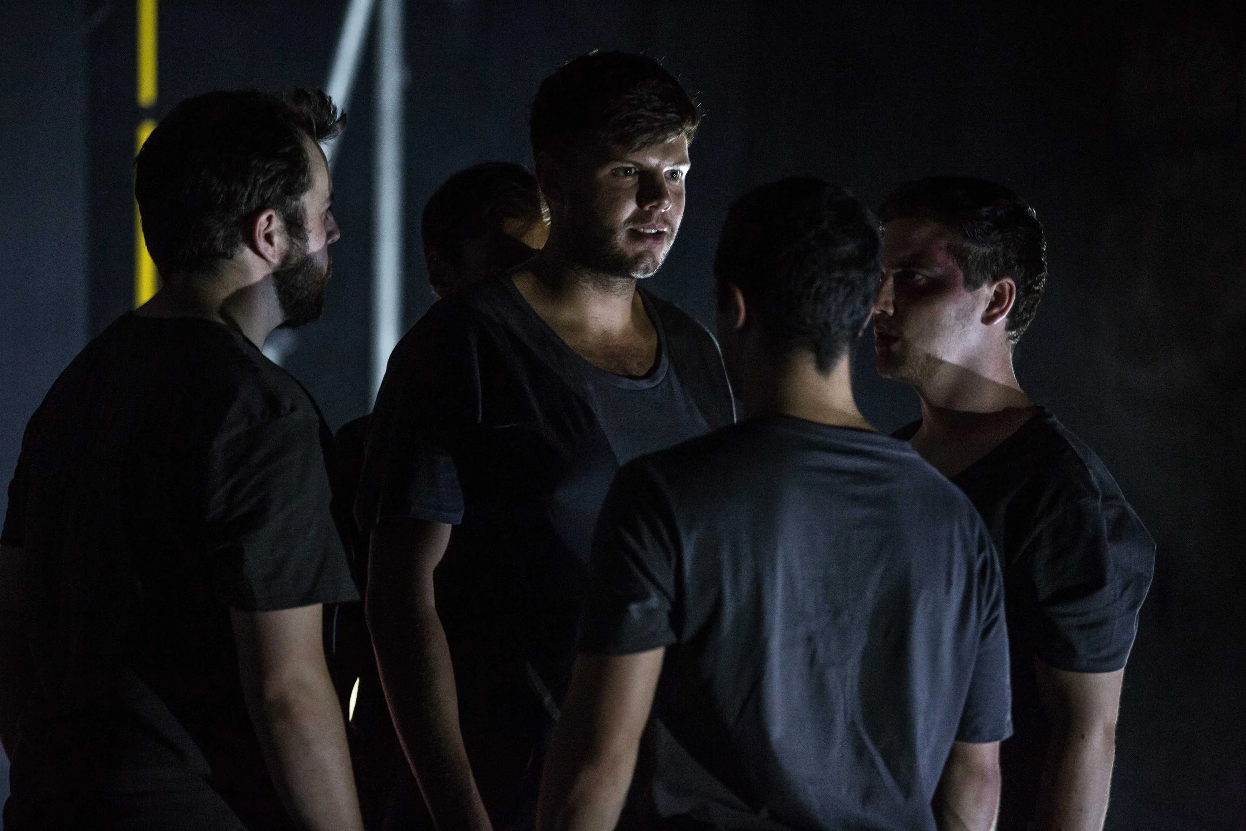 A group of young men standing in a dark room, with some of them looking at each other, illuminated by focused lighting.