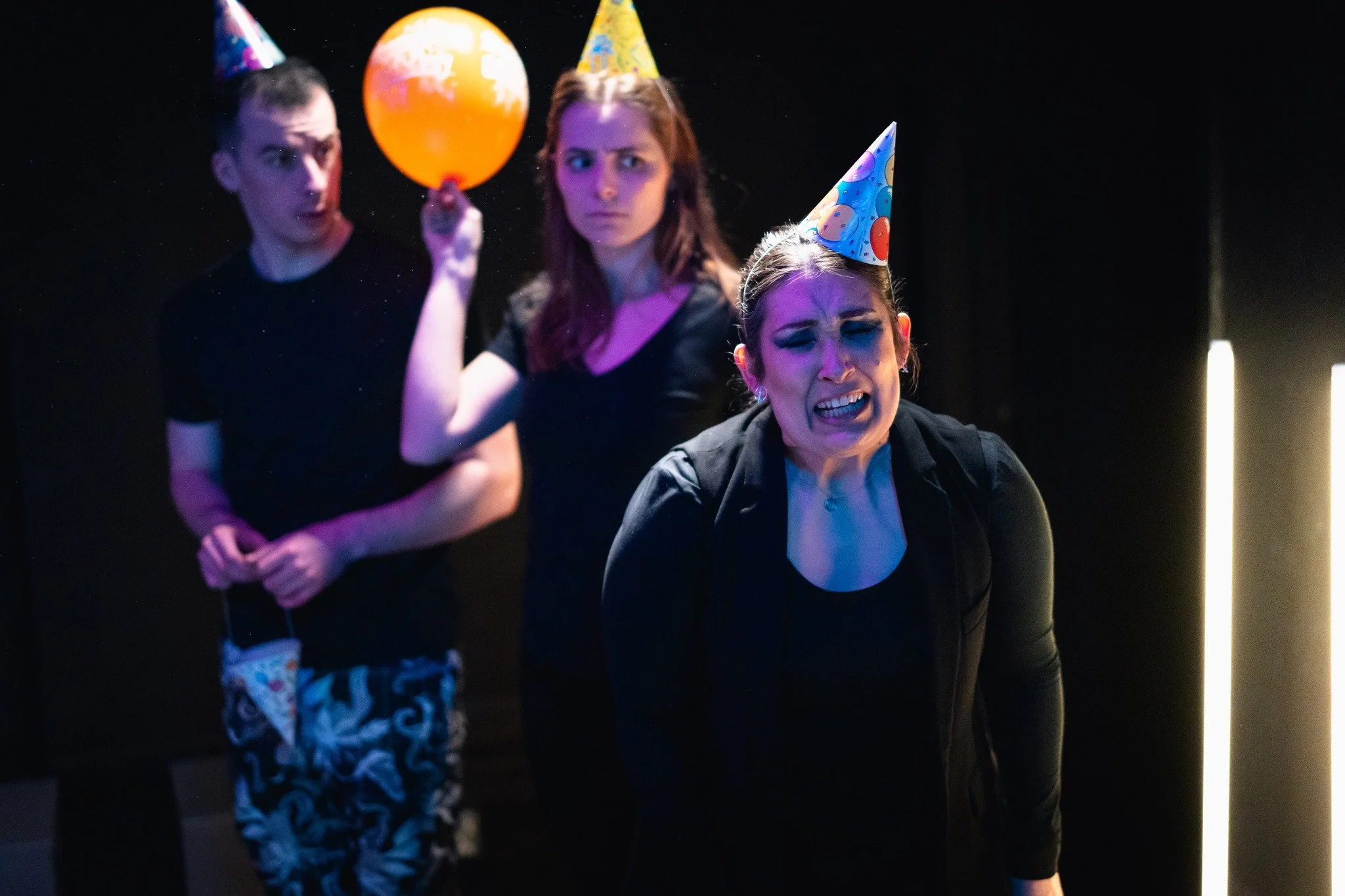 Three people wearing colorful party hats at a celebration, with one person in the foreground making a distressed or confused facial expression and the other two in the background looking on, all against a dark background.