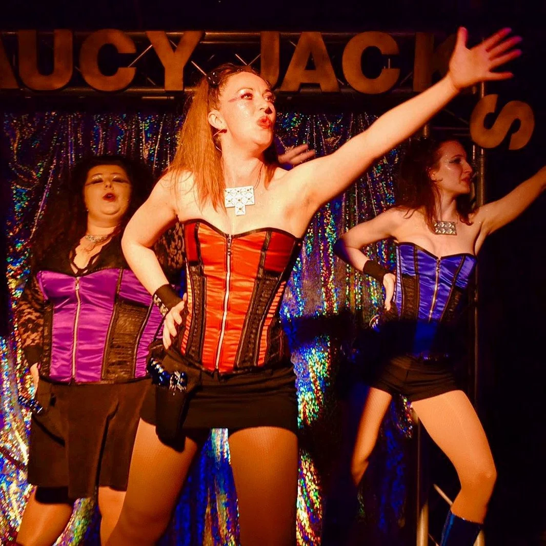 Three women performing on stage with sequined backdrop, dressed in colorful corset tops and shorts, striking dramatic poses.