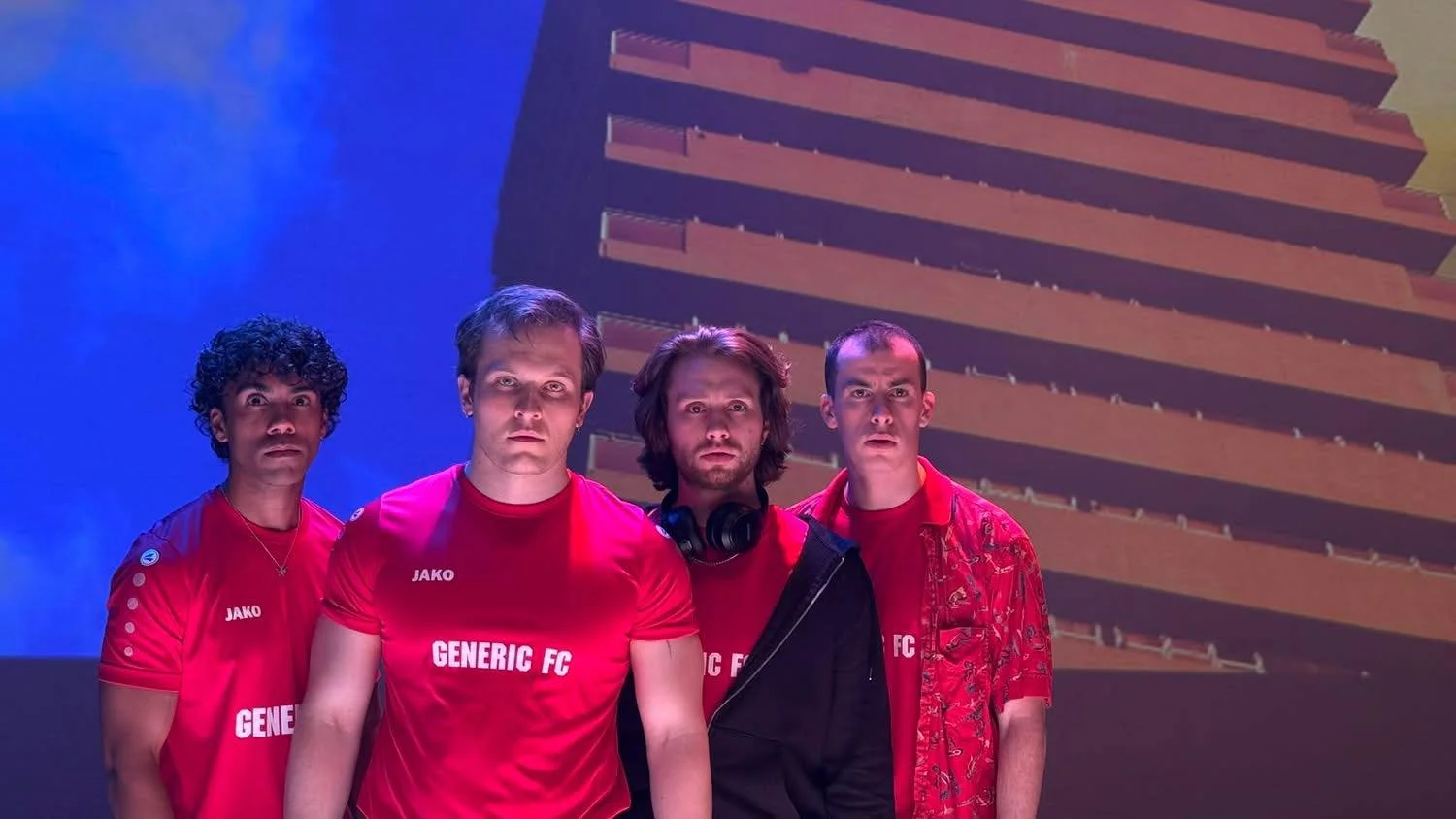 Four men wearing red t-shirts with 'GENERIC FC' and 'JAKO' printed on them, standing in front of a vibrant background with a tall building, one man has headphones around his neck.