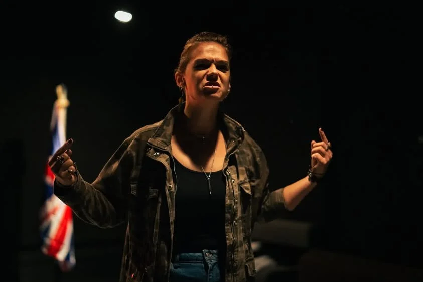 A woman with brown hair pulled back, wearing a camouflage jacket and black top, standing in a dark room with a spotlight above her, making expressive gestures with her hands, and an American flag in the background.