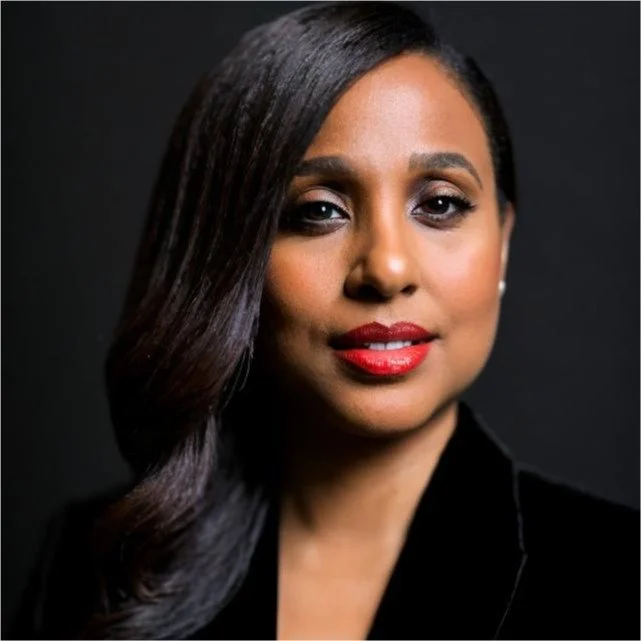 A professional portrait of a woman with long dark hair, wearing red lipstick and a black outfit, against a dark background.