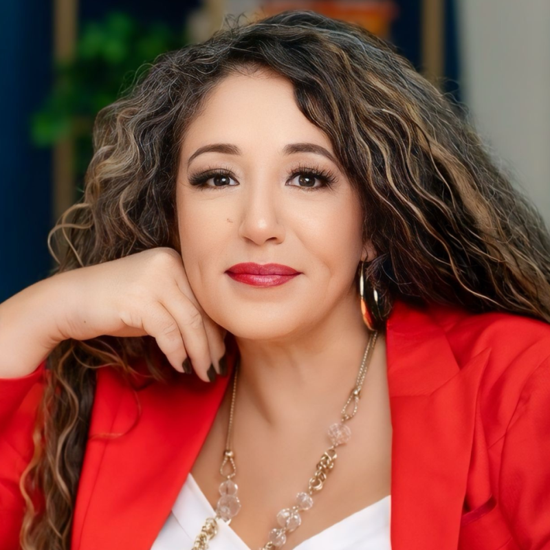 A woman with curly brown hair, wearing a red blazer and statement jewelry, resting her chin on her hand and smiling softly.