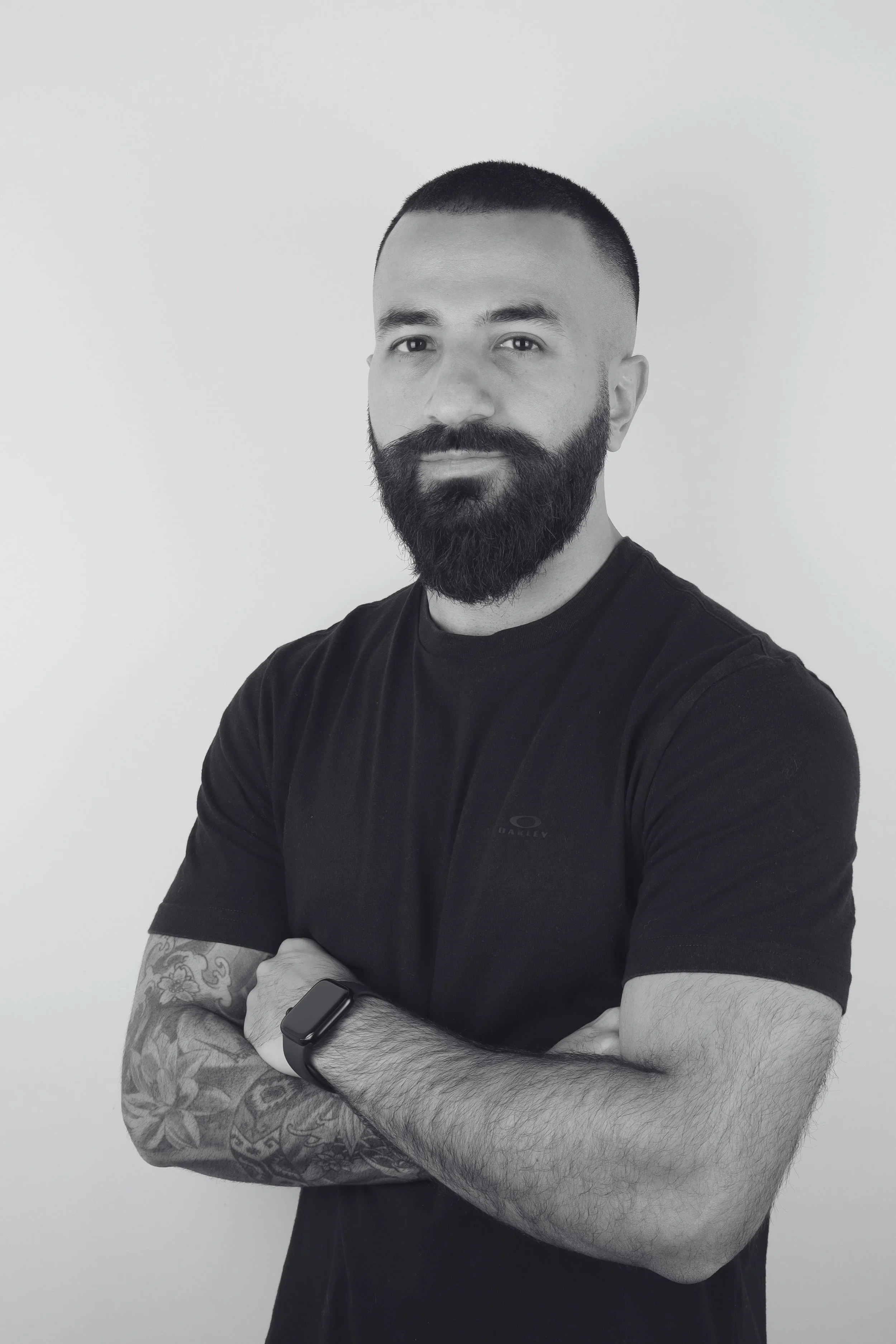 Black and white portrait of a man with a beard and short hair, wearing a black T-shirt with a logo on the chest, standing with crossed arms, against a plain white background.