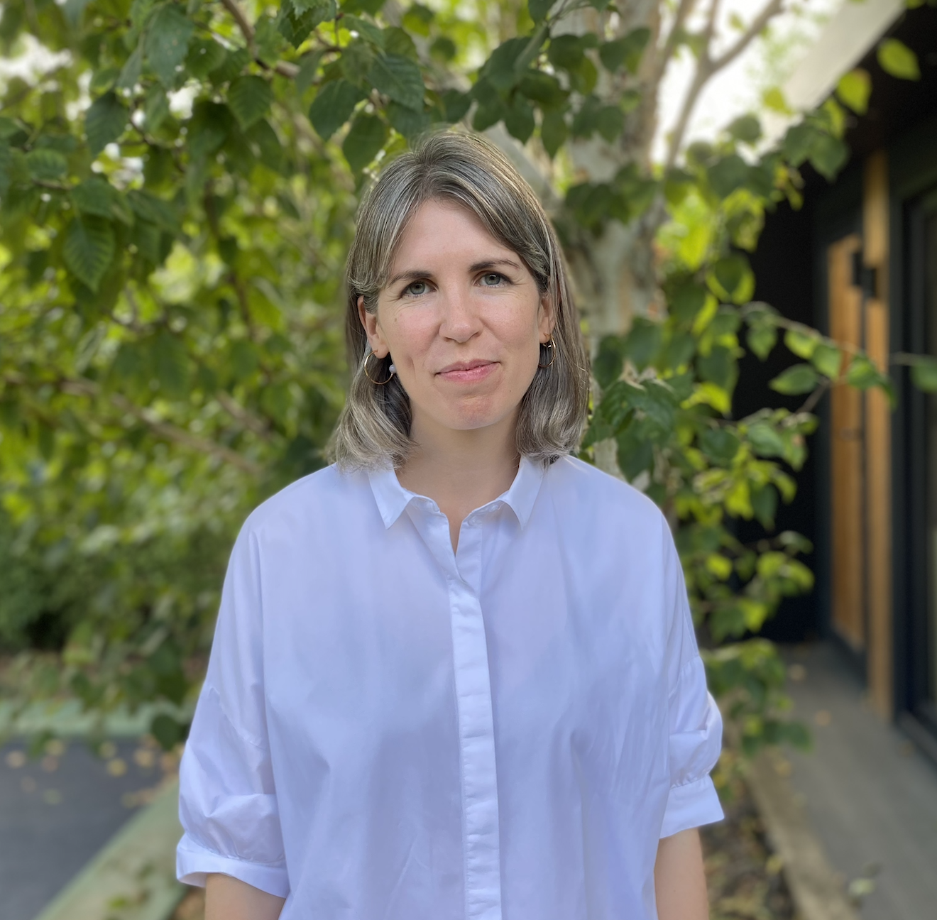 A woman with shoulder-length brown-grey hair and hazel eyes, wearing a white button-up shirt and hoop earrings, standing outdoors in front of green leafy trees and a building with a black exterior and wooden accents.