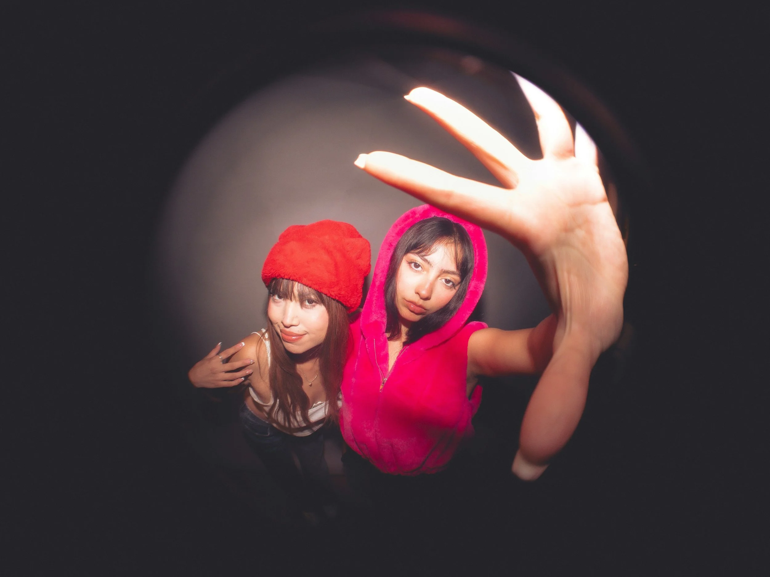 Zwei junge Frauen fotografieren sich selbst in einem Spiegel. Eine trägt einen roten Hut, die andere einen pinken Hoodie.
