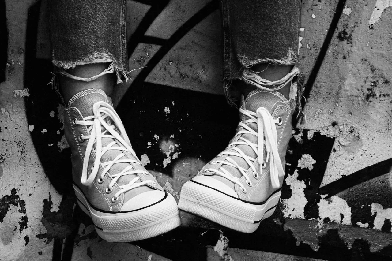 Schwarze und weiße Converse-Sneaker, getragen mit zerrissenen Jeans, auf einem Boden mit Rissen und Schmutz. Das Bild ist in Schwarz-Weiß.