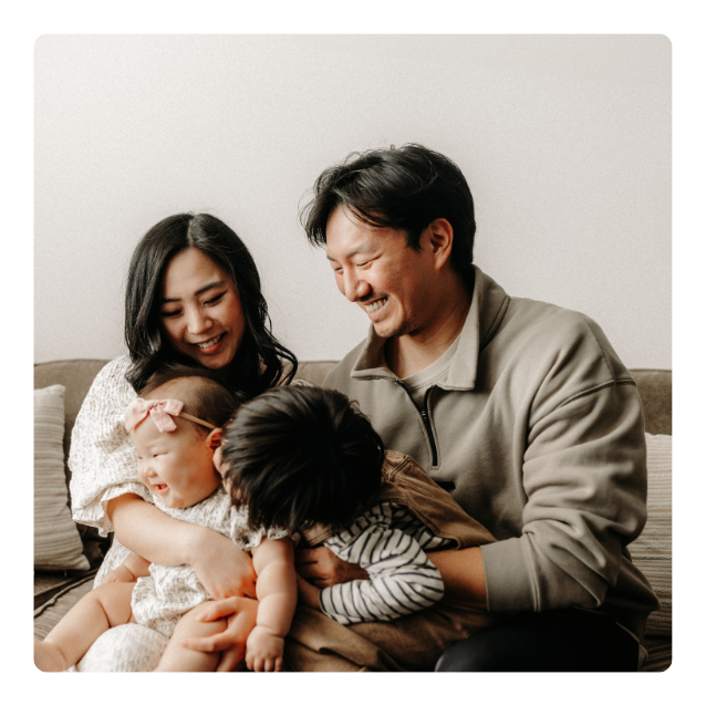 A happy family with two parents and two young children sitting on a couch, smiling and laughing together.