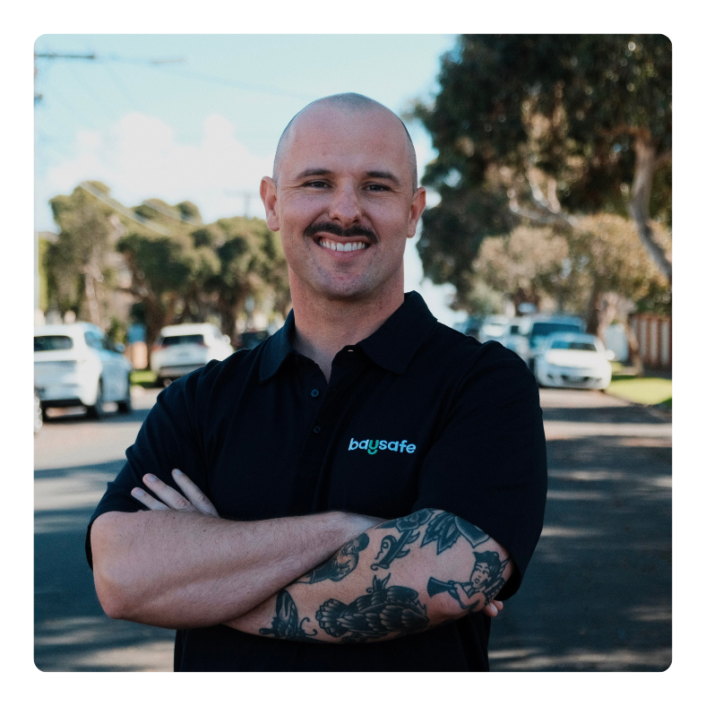 A man with a shaved head and a mustache, wearing a black polo shirt with 'baysafe' logo, standing outdoors with crossed arms, in front of parked cars and trees.
