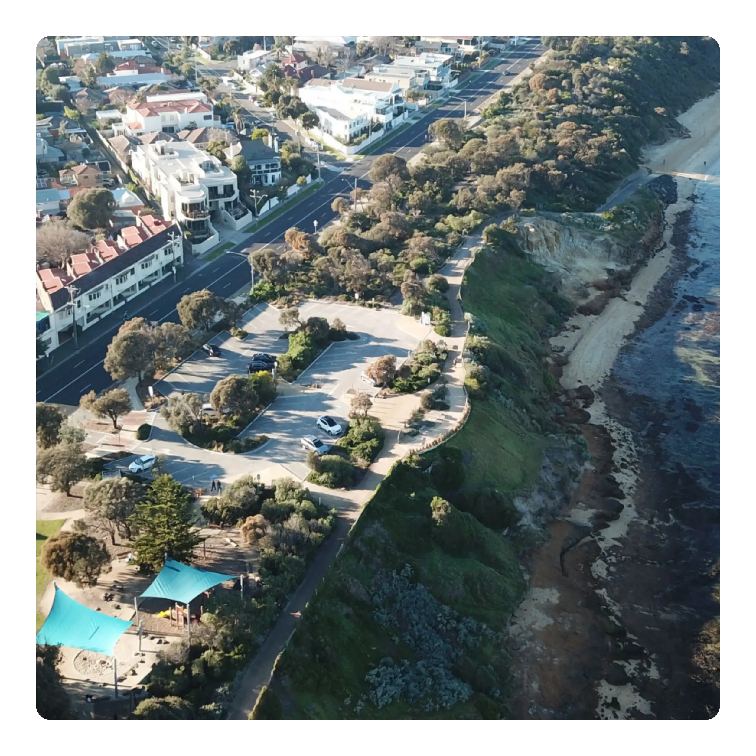 An aerial view of a neighborhood next to a coastline with cliffs, showing residential buildings, trees, and parked cars.