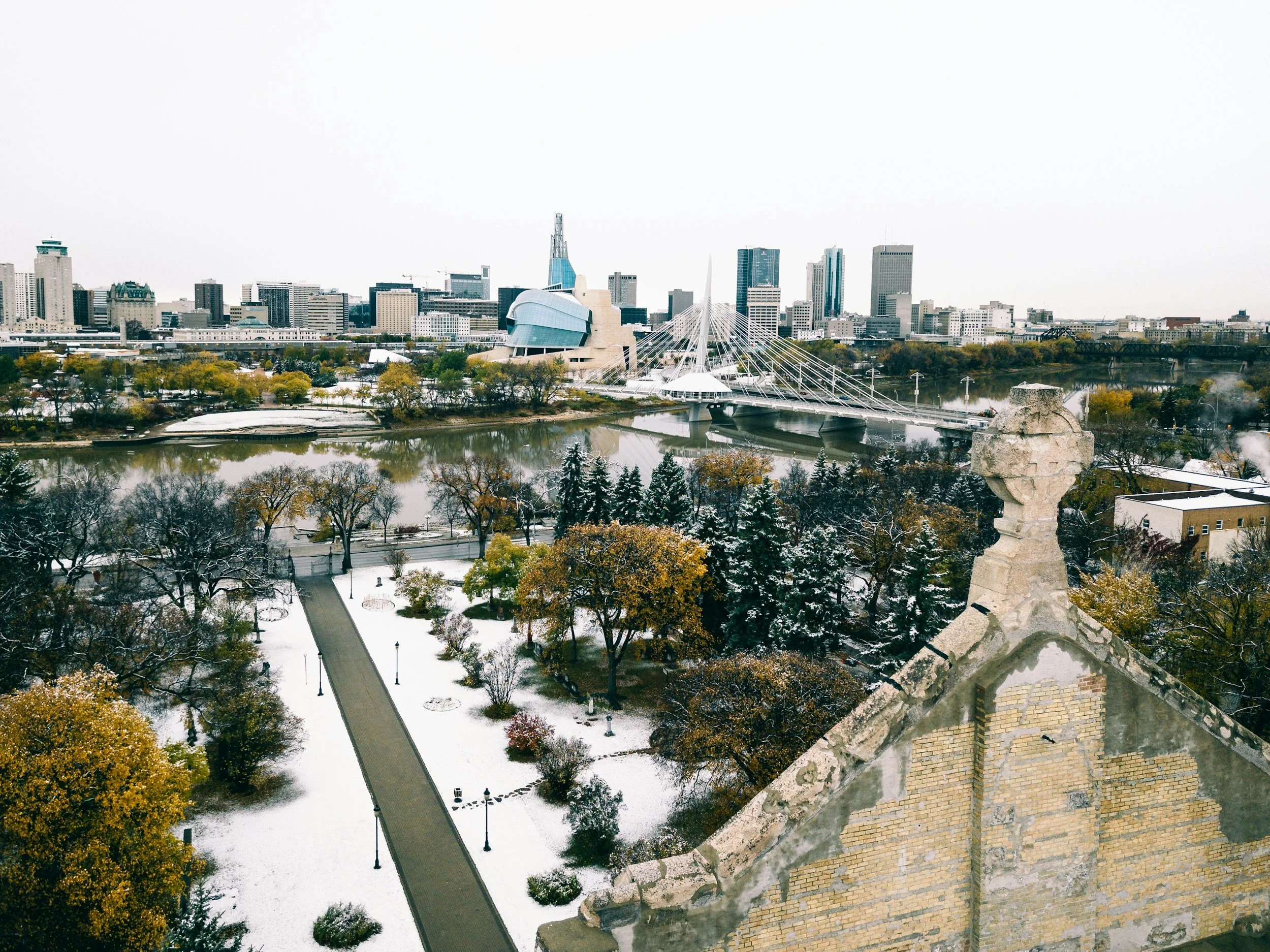 A panoramic cityscape of Winnipeg, Manitoba, Canada, featuring modern skyscrapers, the Canadian Museum for Human Rights with unique architecture, a bridge over the river, and trees with autumn foliage and patches of snow in the foreground.