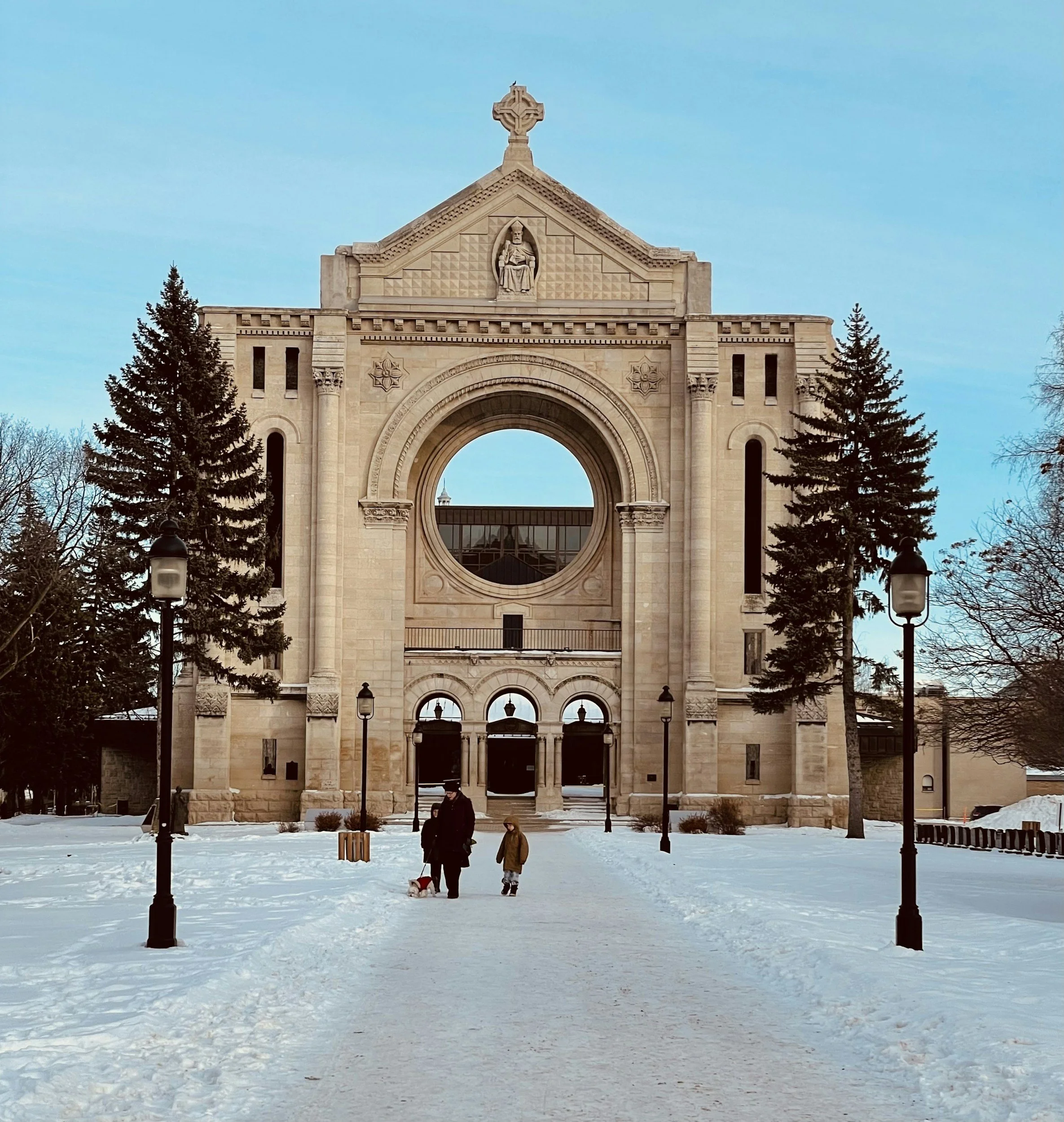 A historic stone archway with a sculpture of a seated figure at the top center, surrounded by trees and flanked by snow-covered ground and street lamps in a park during winter.