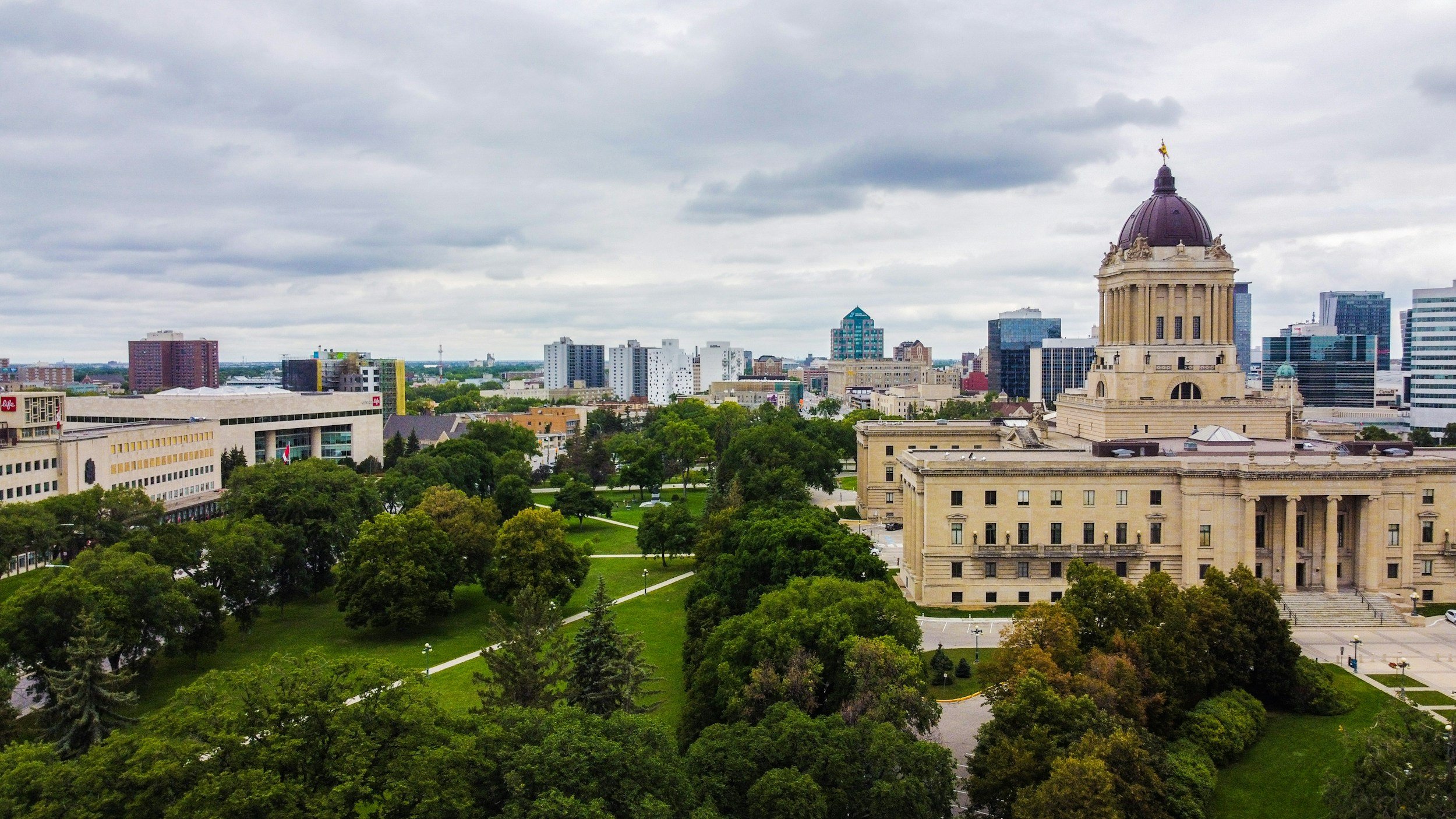 A city skyline with a historic building featuring a dome in the foreground, surrounded by green parks and modern high-rise buildings under a cloudy sky.