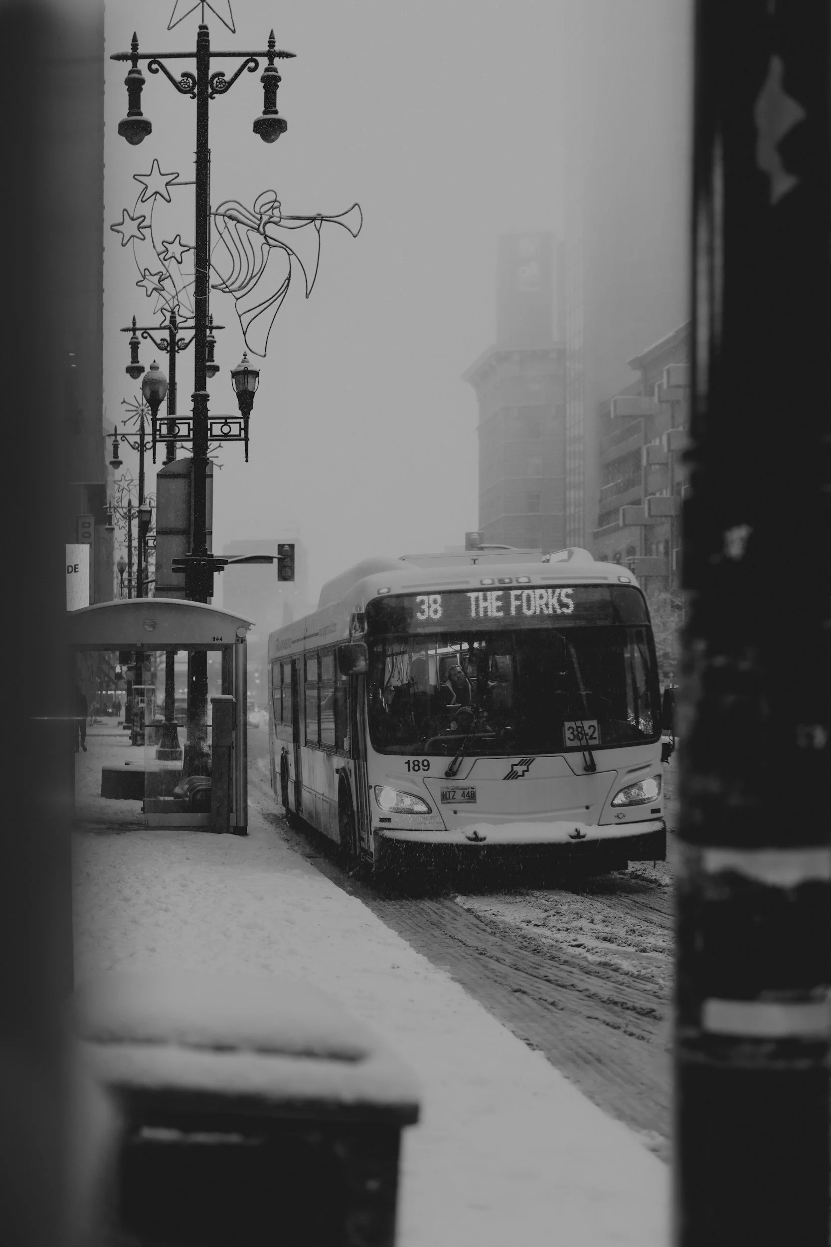 A city scene with a bus labeled 'THE FORKS' approaching a snowy bus stop under decorative streetlights and hanging star-shaped holiday lights, with tall buildings and fog in the background.