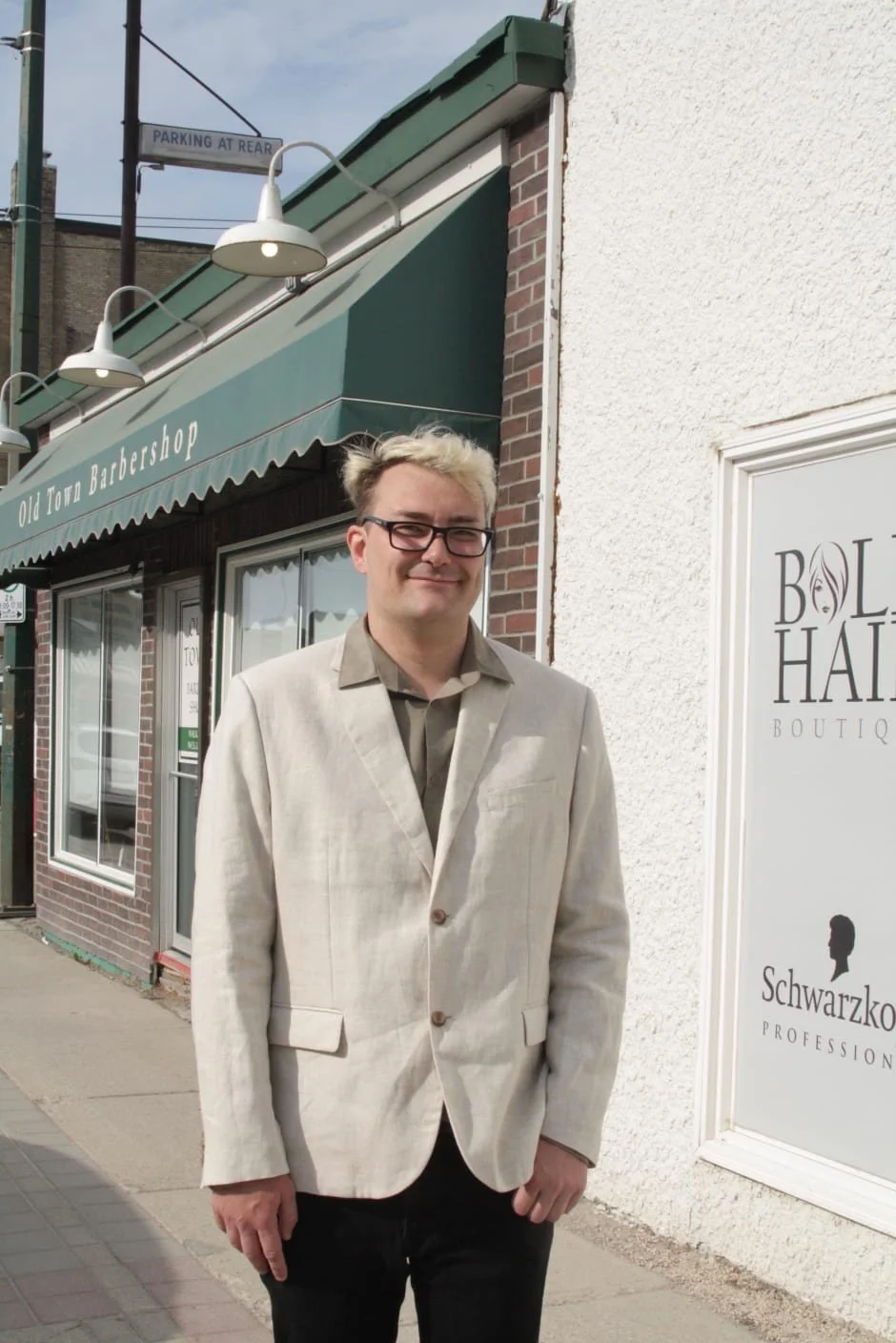 A man with glasses and blonde hair standing outside a storefront with a green awning that reads 'Old Town Barbershop'. He is wearing a beige blazer and a brown shirt, smiling at the camera.